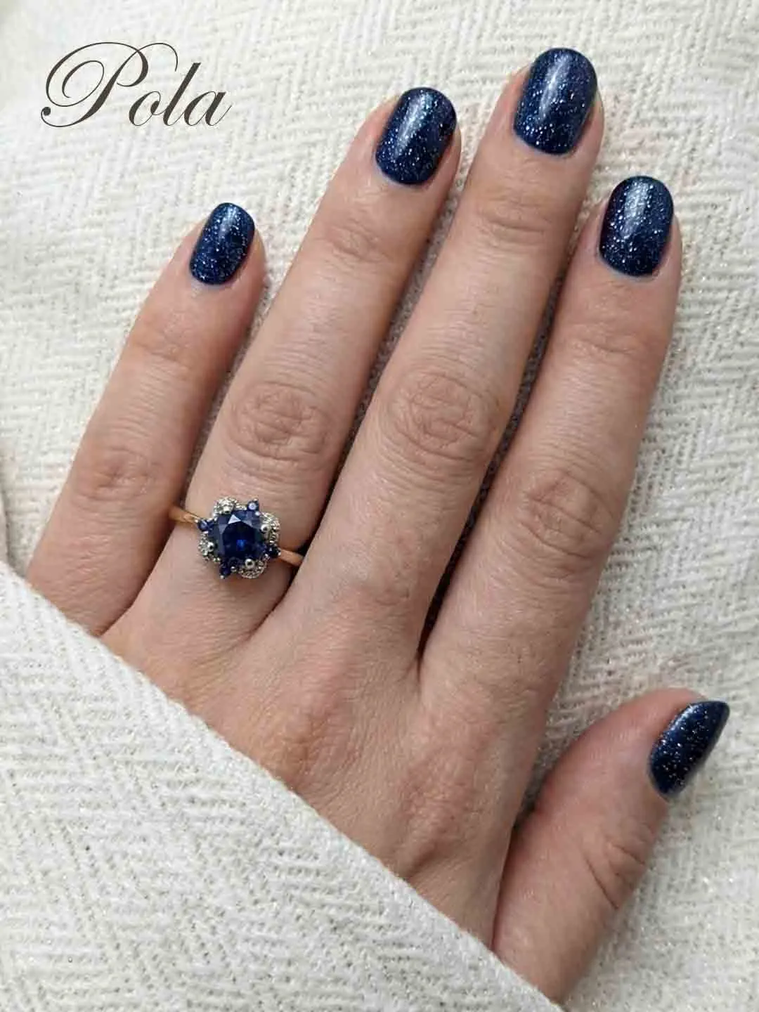 Kobieca dłoń z połyskującym granatowym manicure, prezentująca złoty pierścionek z ciemnoniebieskim kamieniem otoczonym kwiatową aureolą z drobnych cyrkonii.
