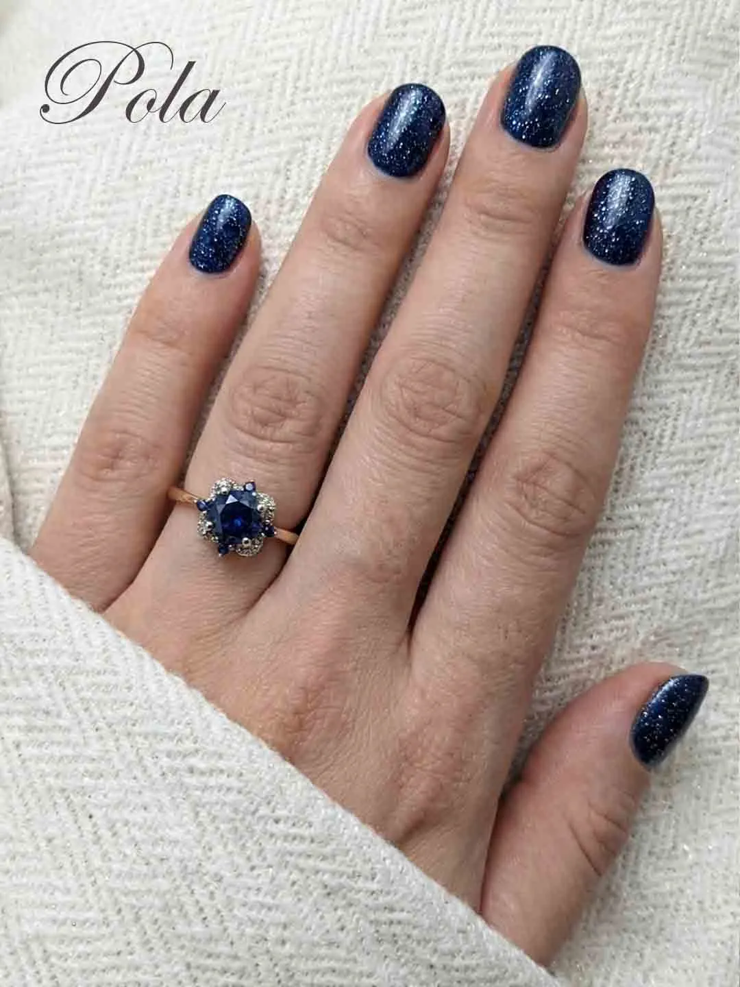 Kobieca dłoń z połyskującym granatowym manicure, prezentująca złoty pierścionek z ciemnoniebieskim kamieniem otoczonym kwiatową aureolą z drobnych cyrkonii.