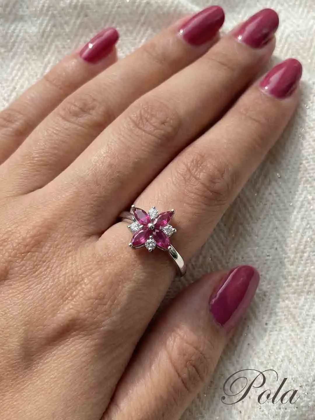 Kobieca dłoń z malinowym manicure, zdobiona srebrnym pierścionkiem przypominającym kwiat, z różowymi kamieniami w szlifie markizy i lśniącymi detalami.