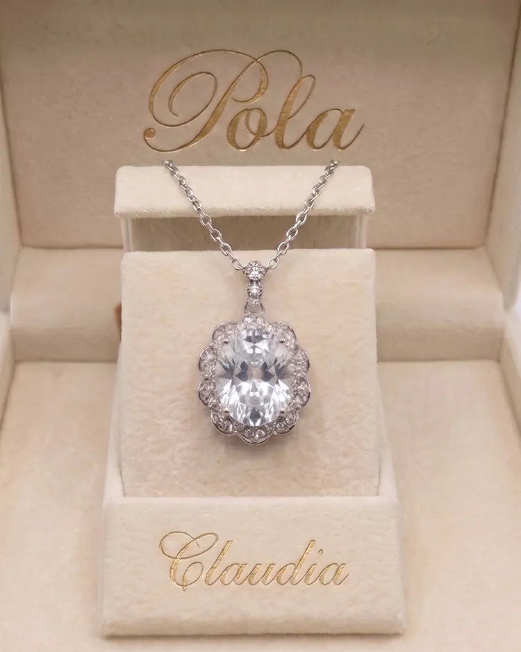 Elegancki wisiorek na łańcuszku ze srebra rodowanego z dużym, owalnym bezbarwnym szafirem, otoczonym drobnymi kamieniami, ułożony w kremowym pudełku jubilerskim marki Pola.